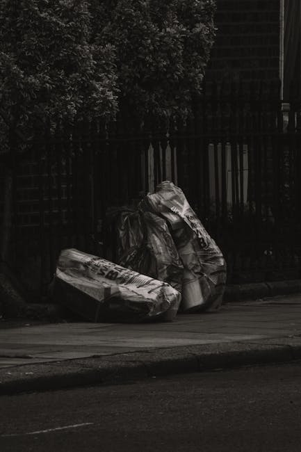 Three large clear plastic bags filled with waste are positioned along the curbside adjacent to a black metal fence and a brick wall, with a section of pavement and a small amount of road visible in th