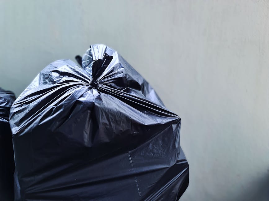 Three large clear plastic bags filled with waste are positioned along the curbside adjacent to a black metal fence and a brick wall, with a section of pavement and a small amount of road visible in th